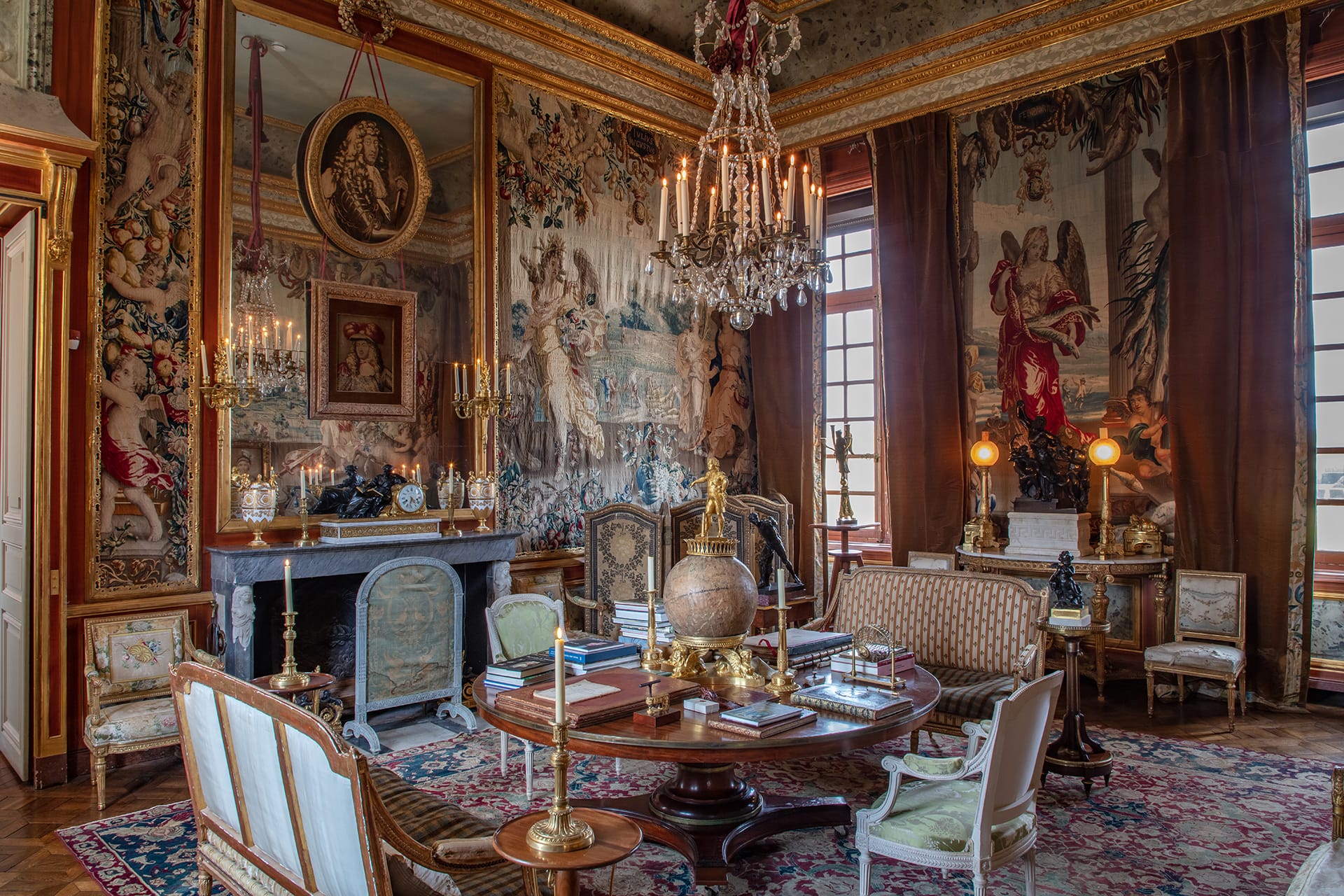 Lavish historical sitting room with ornate furniture, large tapestry, chandelier, fireplace, and globe on a central table.