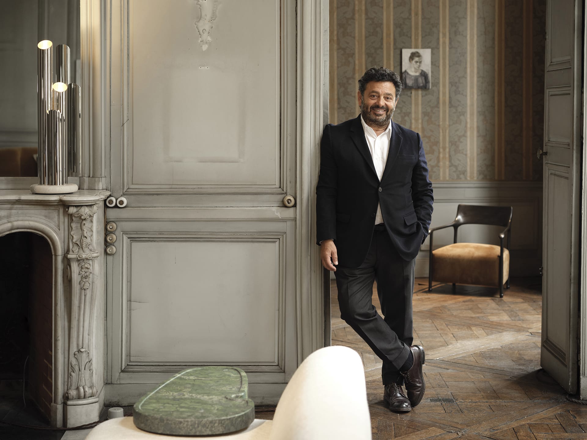 Man in a suit standing in an elegant room with wood floors, next to a fireplace and modern furniture.