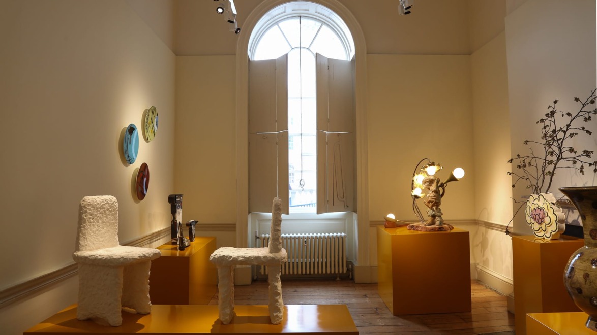 Art gallery interior featuring sculptural chairs, decorative plates on the wall, and elegant lighting near a large arched window.