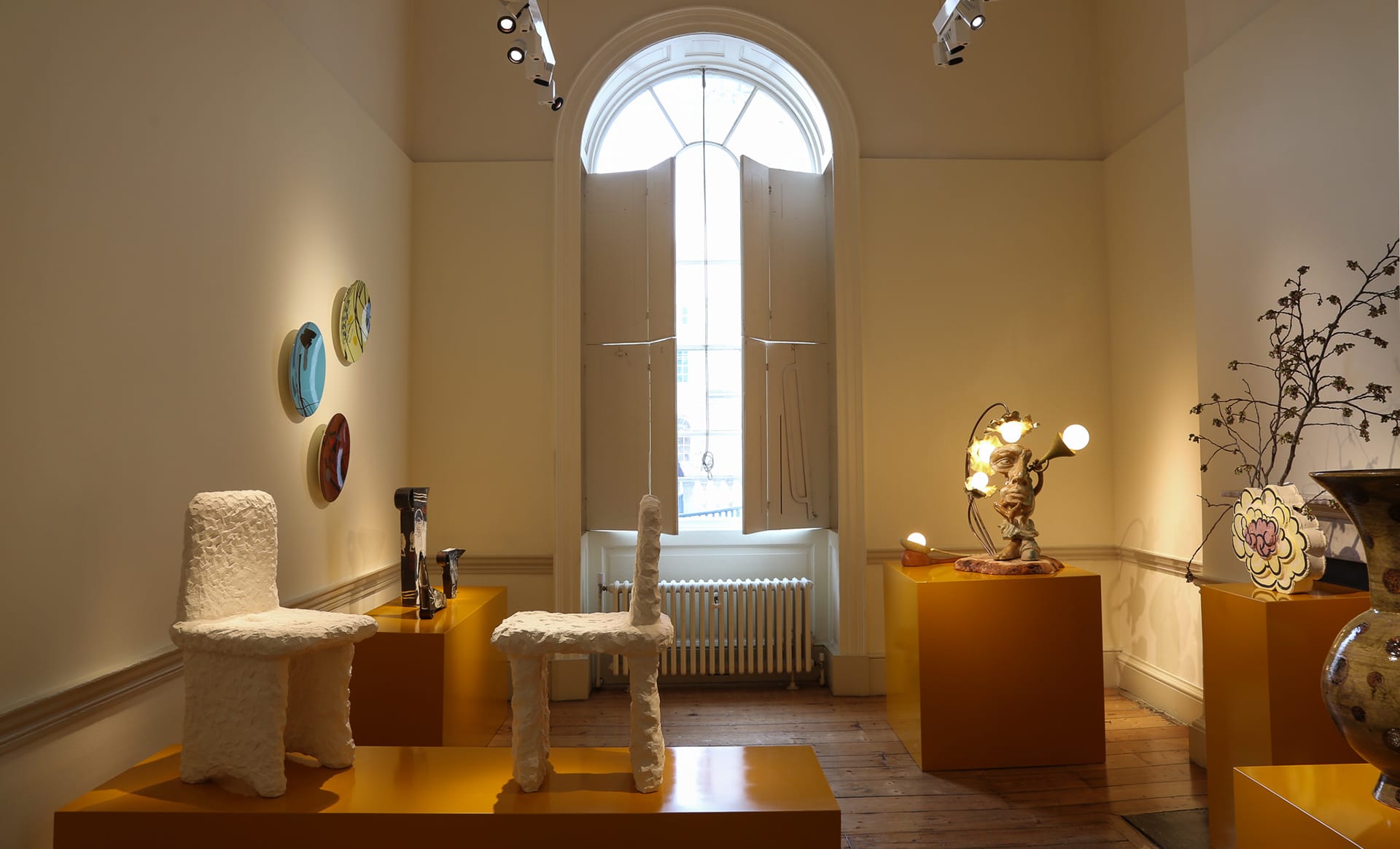 Art gallery interior featuring sculptural chairs, decorative plates on the wall, and elegant lighting near a large arched window.