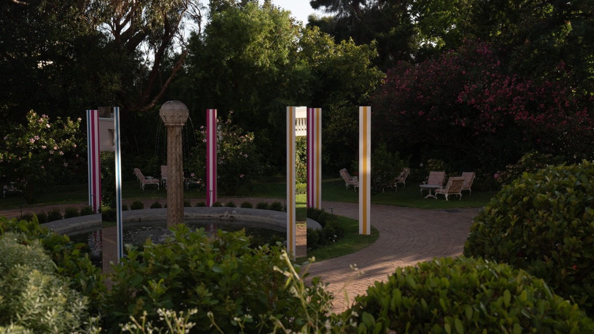 Colorful pillars surround a stone fountain in a lush garden with trees and pathways in the background.