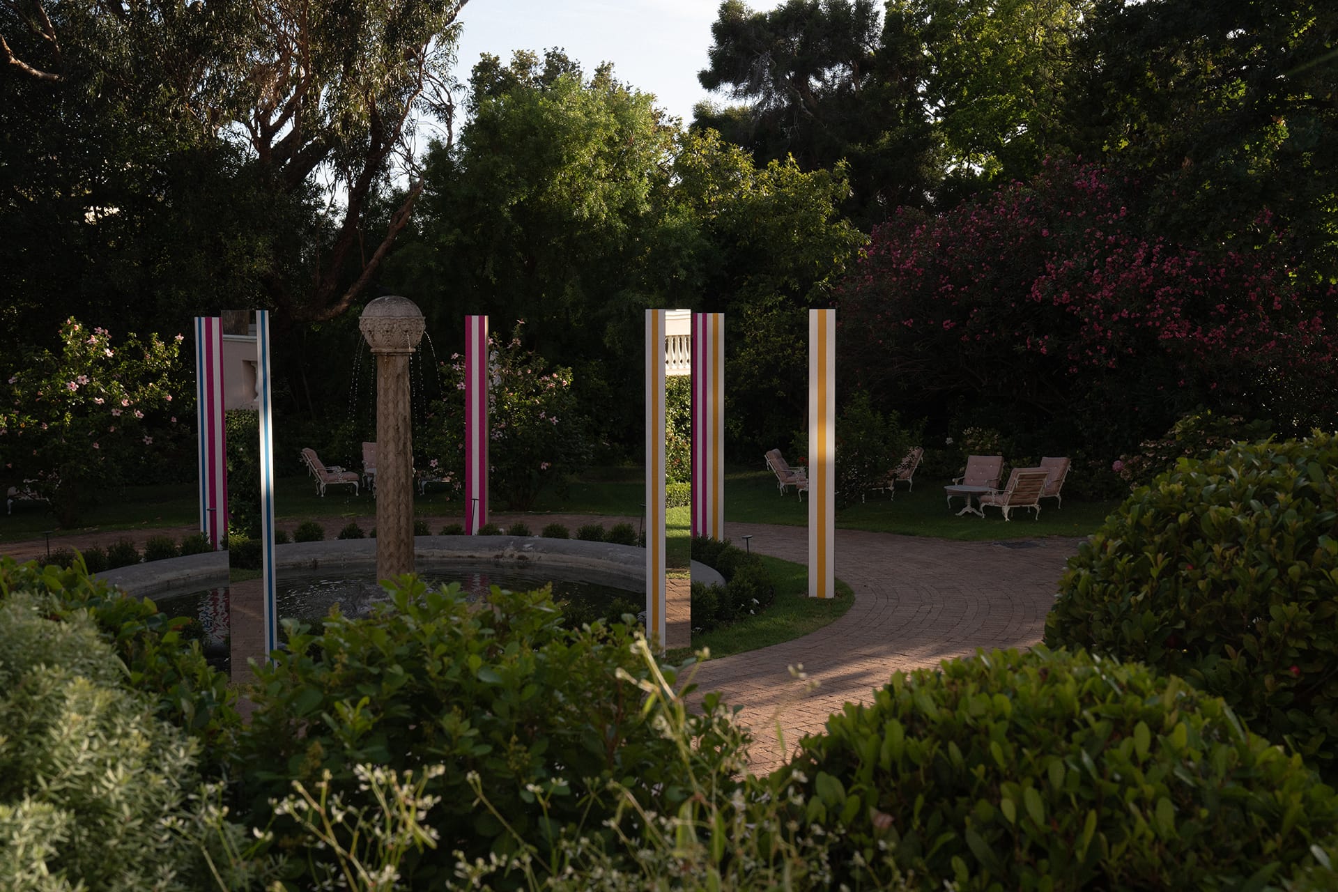 Colorful pillars surround a stone fountain in a lush garden with trees and pathways in the background.