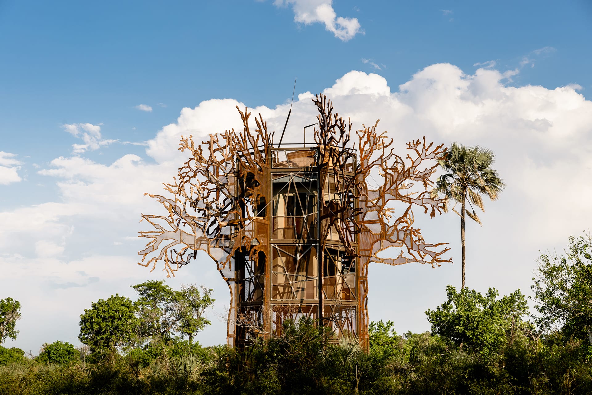 Observation tower surrounded by metal tree sculpture and lush greenery under a blue sky with clouds.
