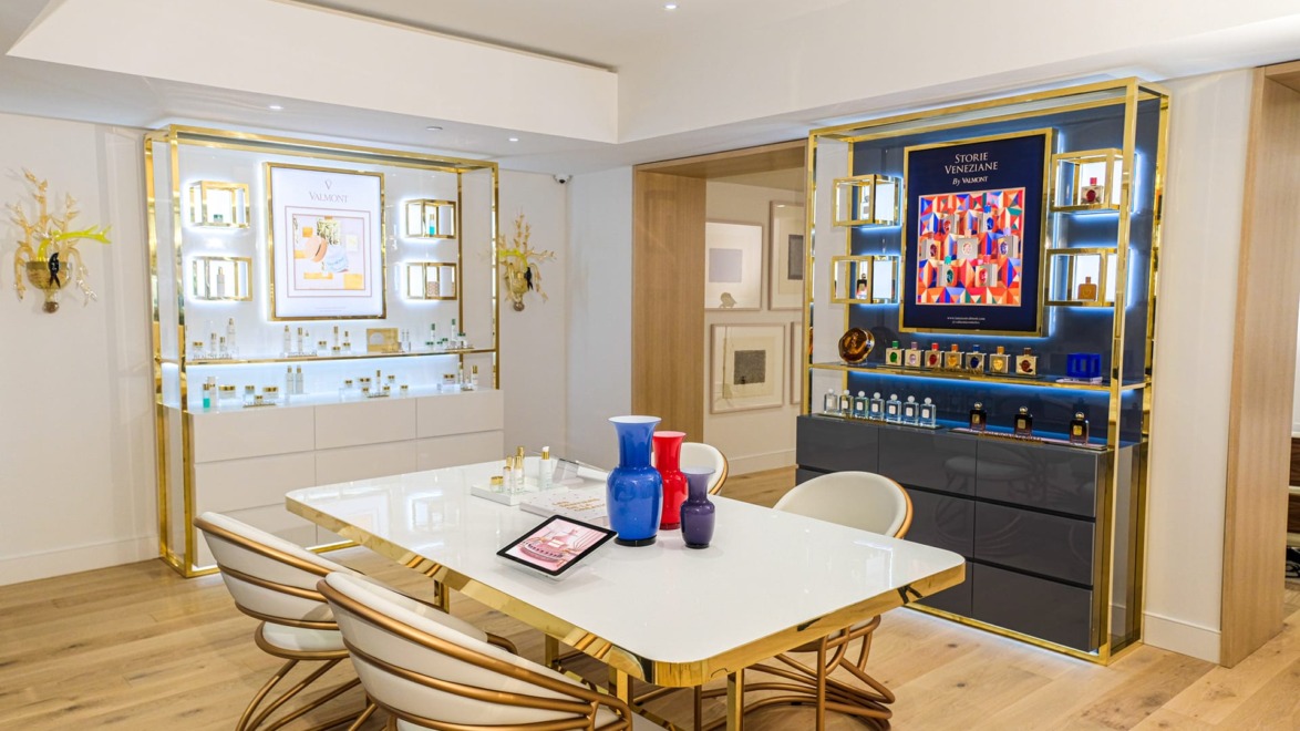 Luxury perfume showroom with gold accents, white table with colorful vases, and shelves displaying various perfume bottles.