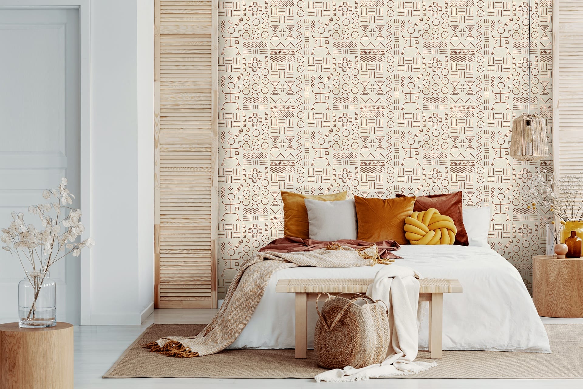Boho-style bedroom with patterned wallpaper, cozy bed, assorted pillows, wooden accents, and woven basket on a bench.