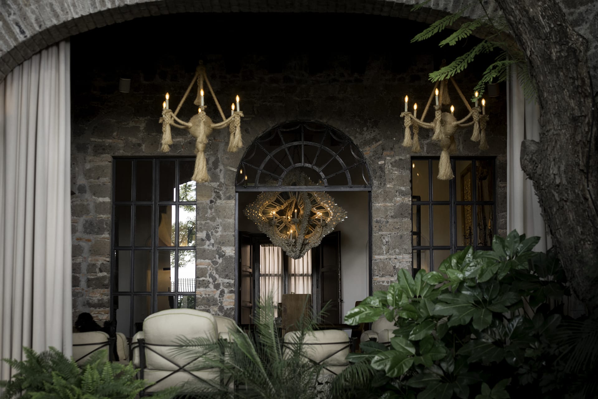 Elegant stone archway reveals a stylish interior with modern chandeliers and plush seating, surrounded by lush greenery.
