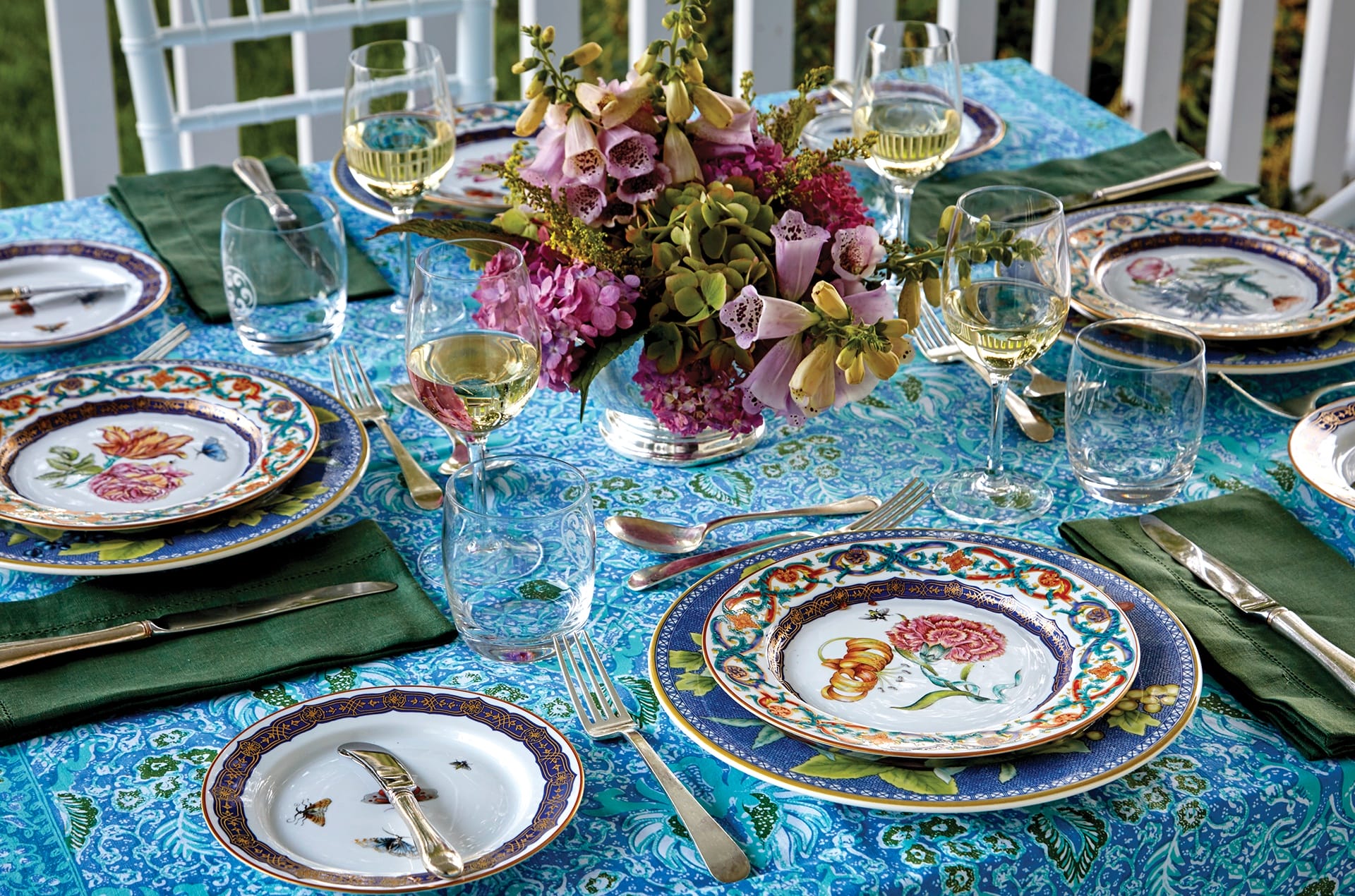 Elegant outdoor table setting with floral china plates, wine glasses, and a floral centerpiece on a blue patterned tablecloth.