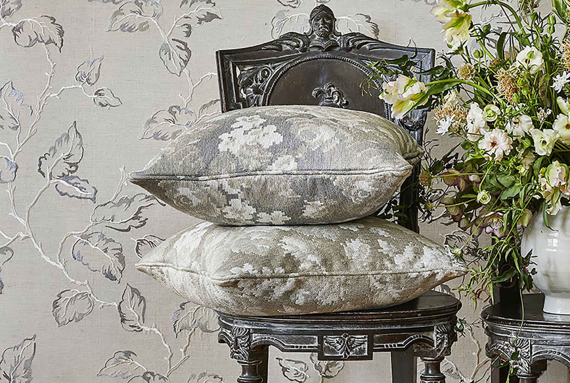 Two decorative pillows on ornate chair beside floral arrangement against a patterned wallpaper background.