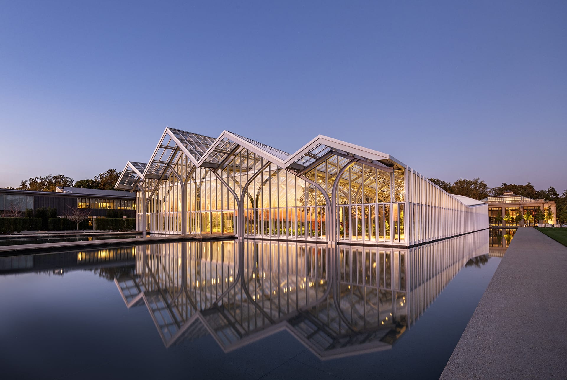 Modern glass building with geometric roof design reflects on calm water during twilight, surrounded by trees and soft evening light.