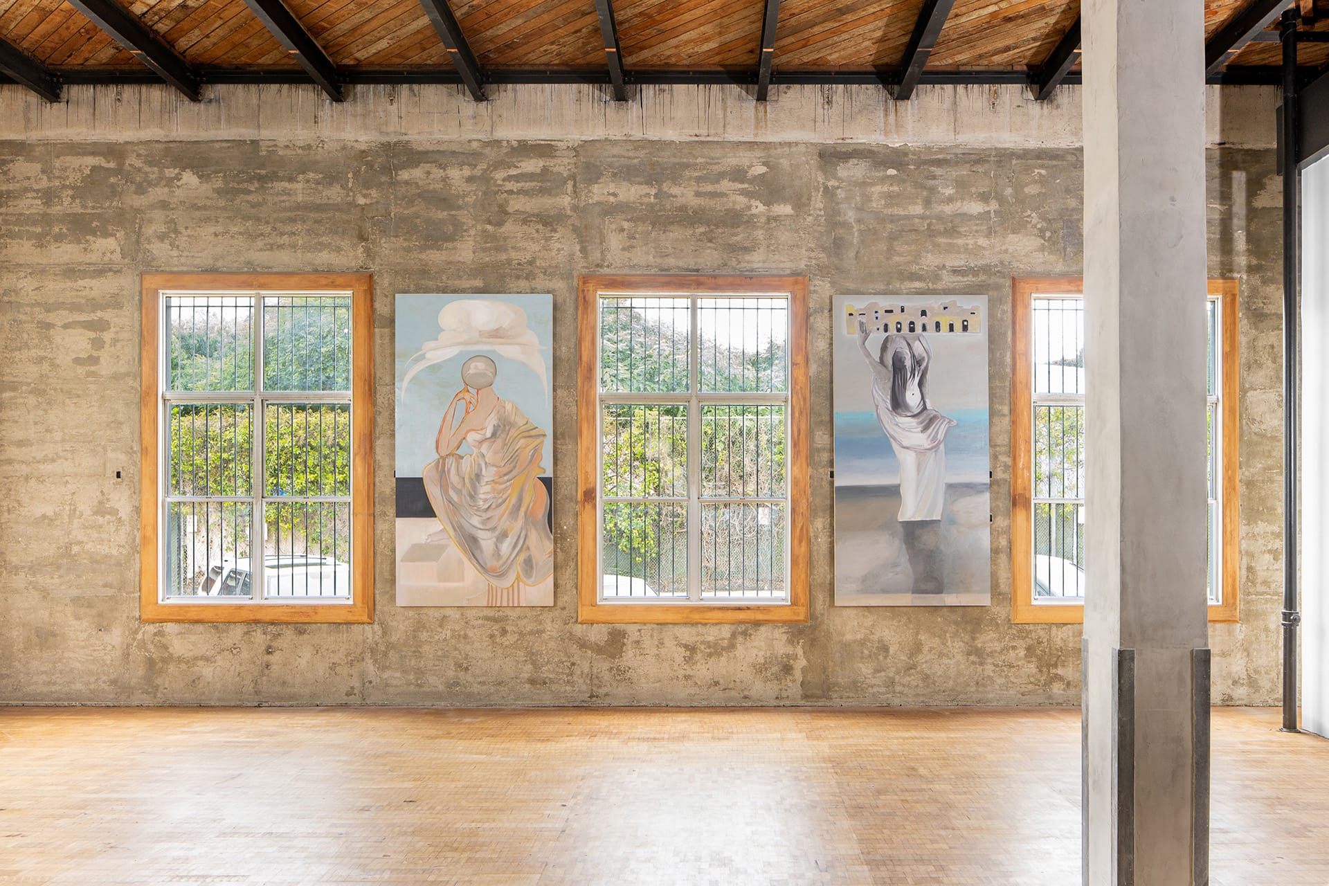 Three large paintings hanging on a rough concrete wall between windows with green metal bars in an industrial art gallery.