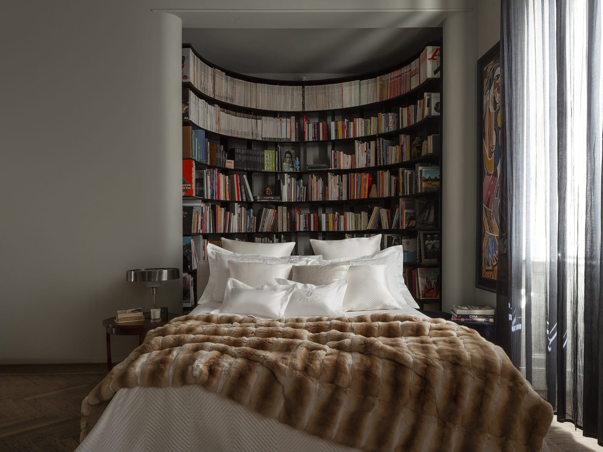 Cozy bedroom with a fur throw blanket on the bed, surrounded by a curved bookshelf filled with books and art decor.