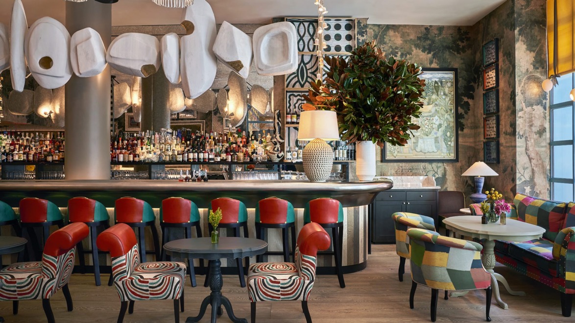 Colorful and eclectic bar interior with vibrant chairs, wooden floor, large plant, and modern decorative elements.