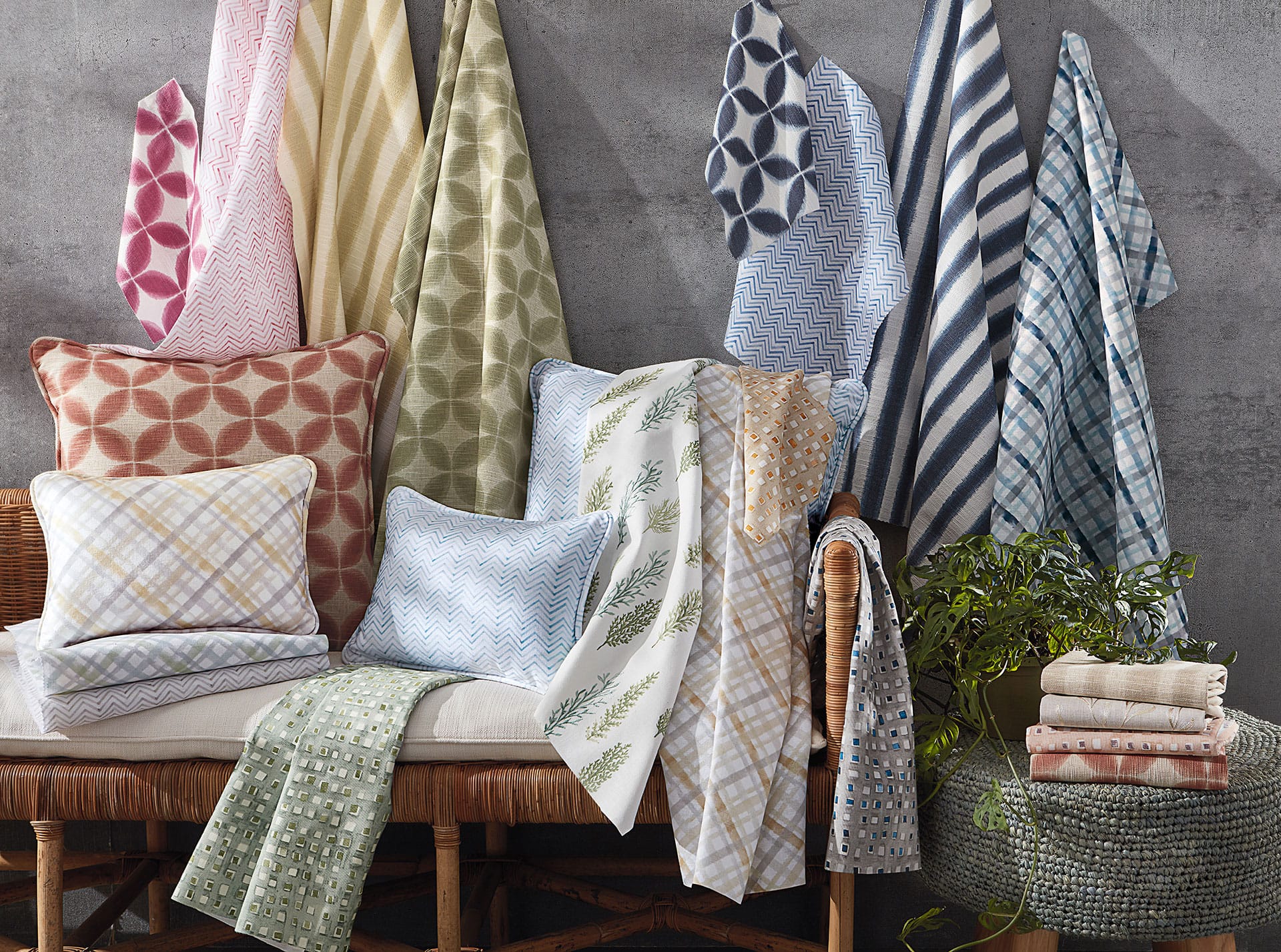 Assorted colorful patterned blankets and pillows displayed on a bench with a plant and towels nearby.