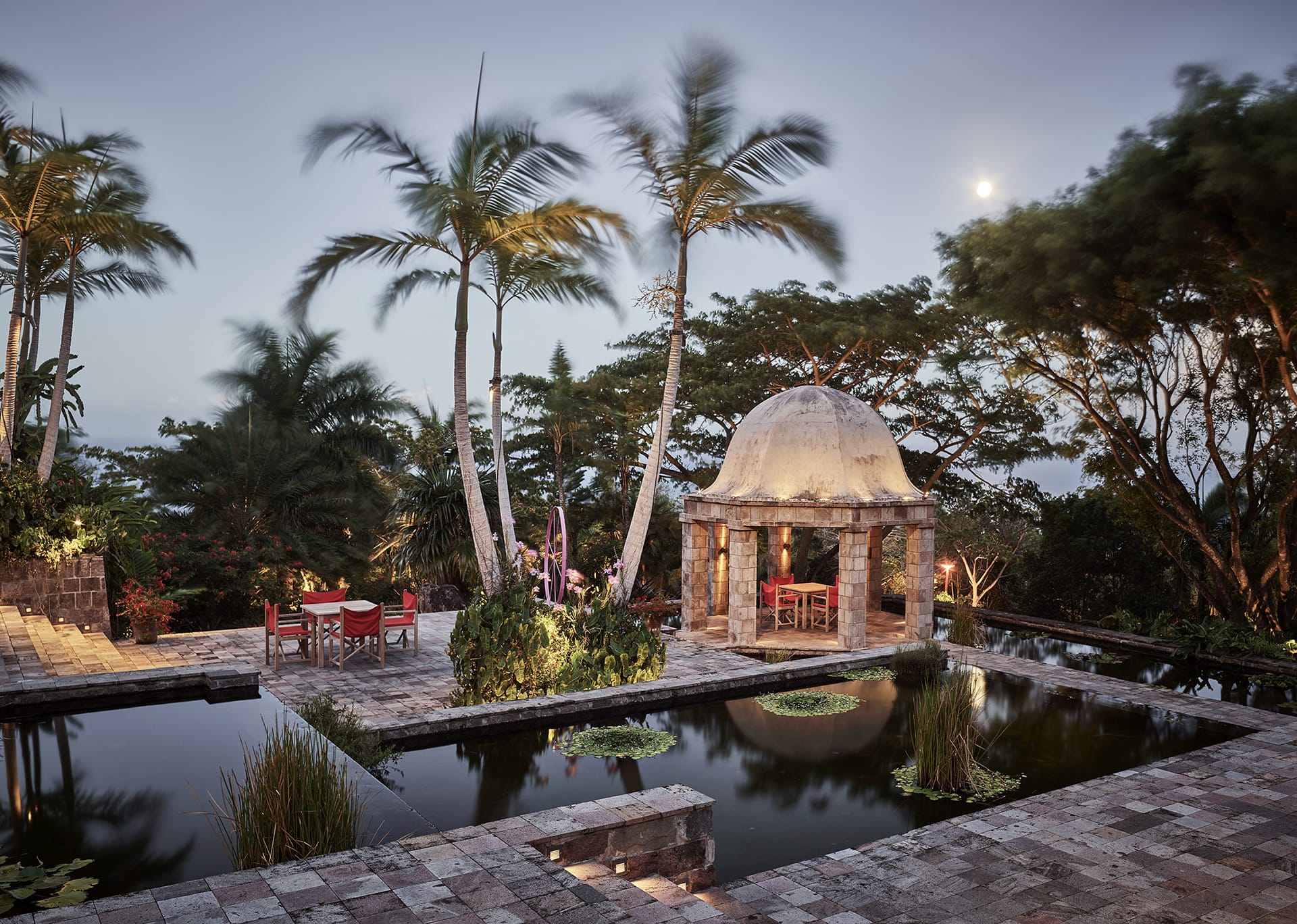 Tropical outdoor setting with stone pavilion, palm trees, and reflective pond at dusk.