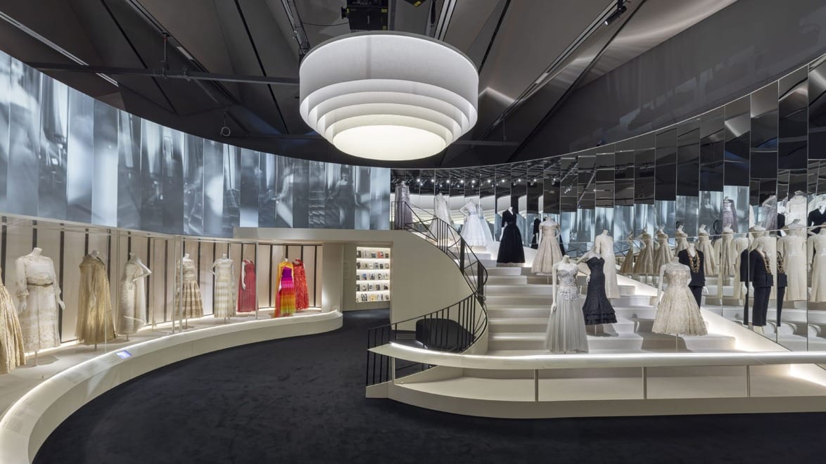 Exhibit of elegant dresses displayed on mannequins in a modern museum setting with a spiral staircase.