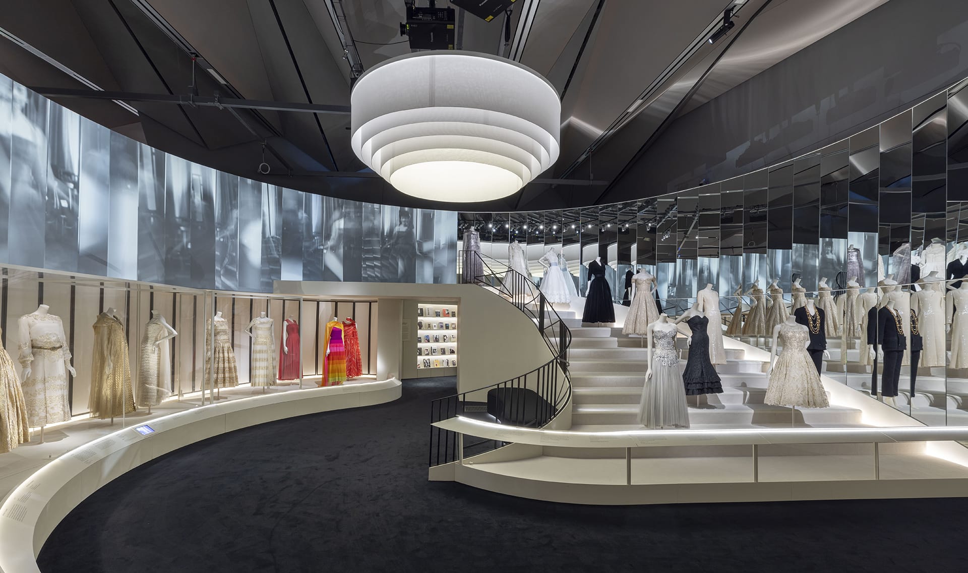 Exhibit of elegant dresses displayed on mannequins in a modern museum setting with a spiral staircase.