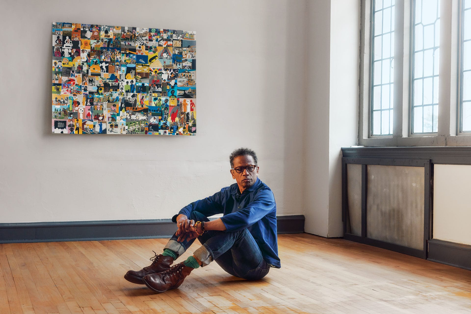 Man sitting on wooden floor in art gallery, colorful collage artwork on wall, large windows letting in natural light.
