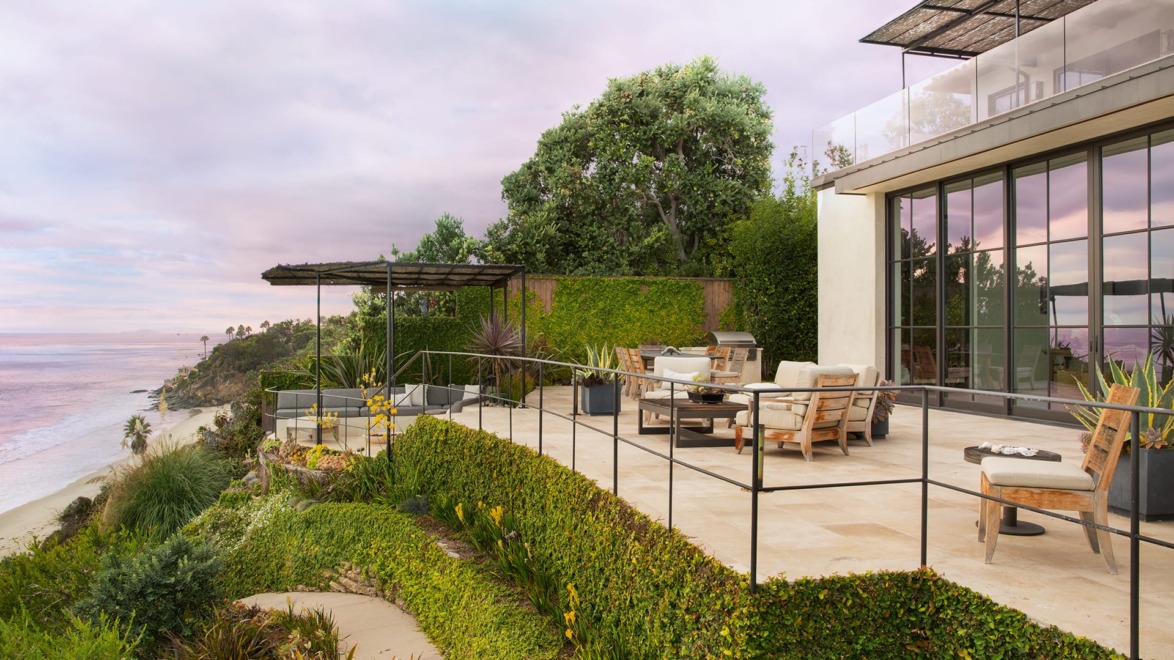Oceanfront patio with modern outdoor furniture overlooking a beach, surrounded by lush greenery and a colorful sky.