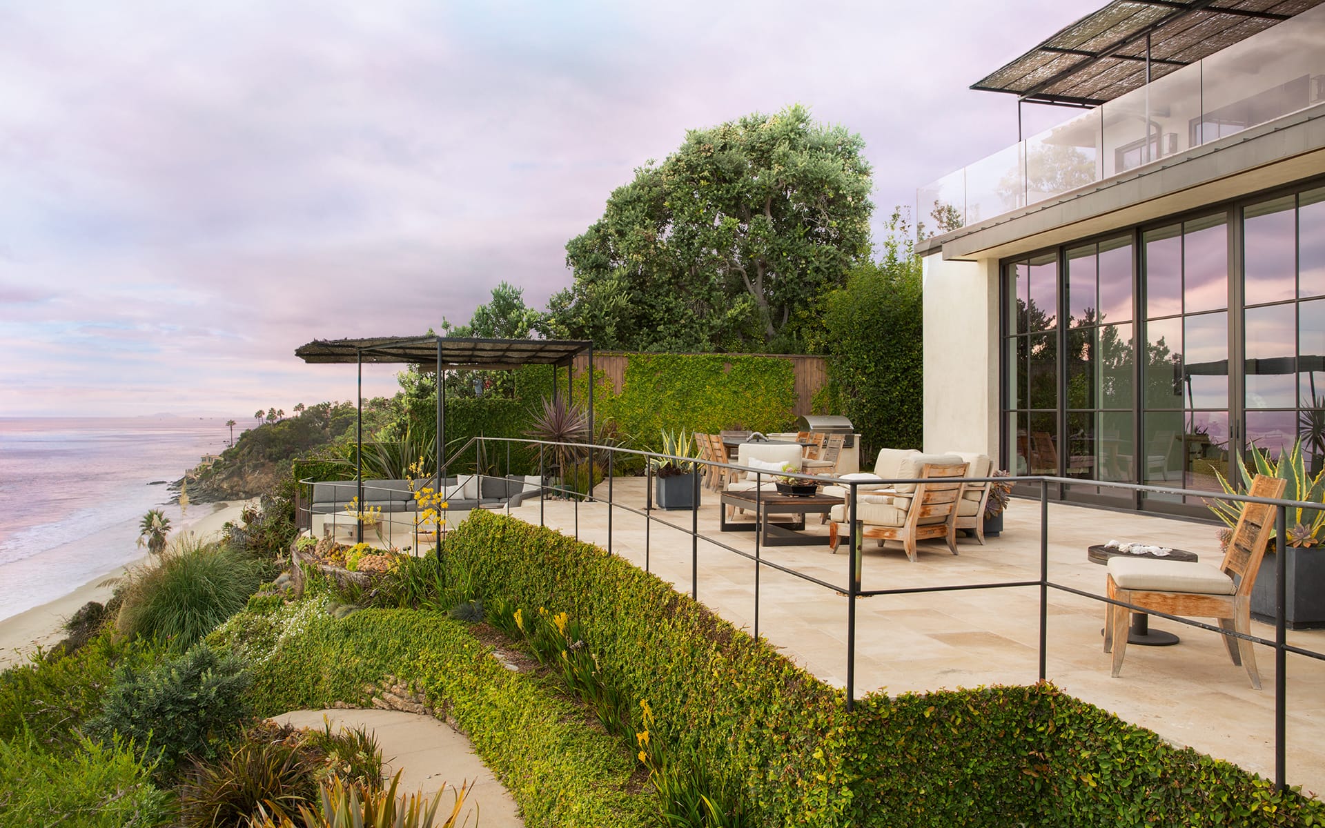Oceanfront patio with modern outdoor furniture overlooking a beach, surrounded by lush greenery and a colorful sky.