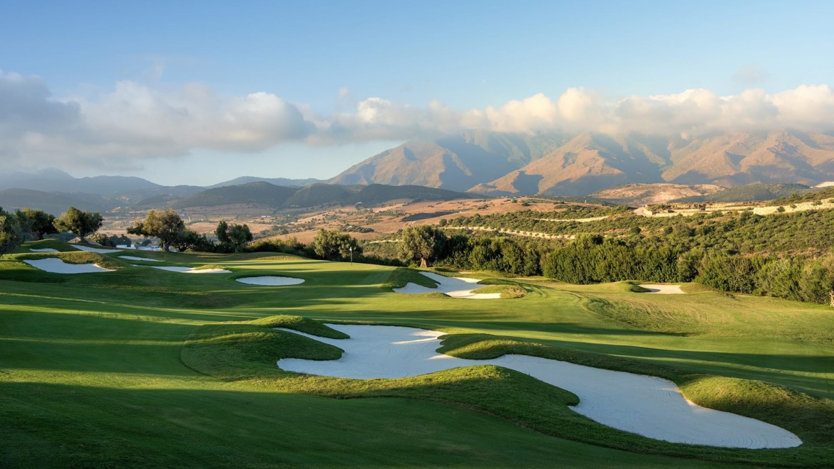 Scenic view of a golf course with sand traps and lush greenery, backdrop of mountains under a blue sky with clouds.