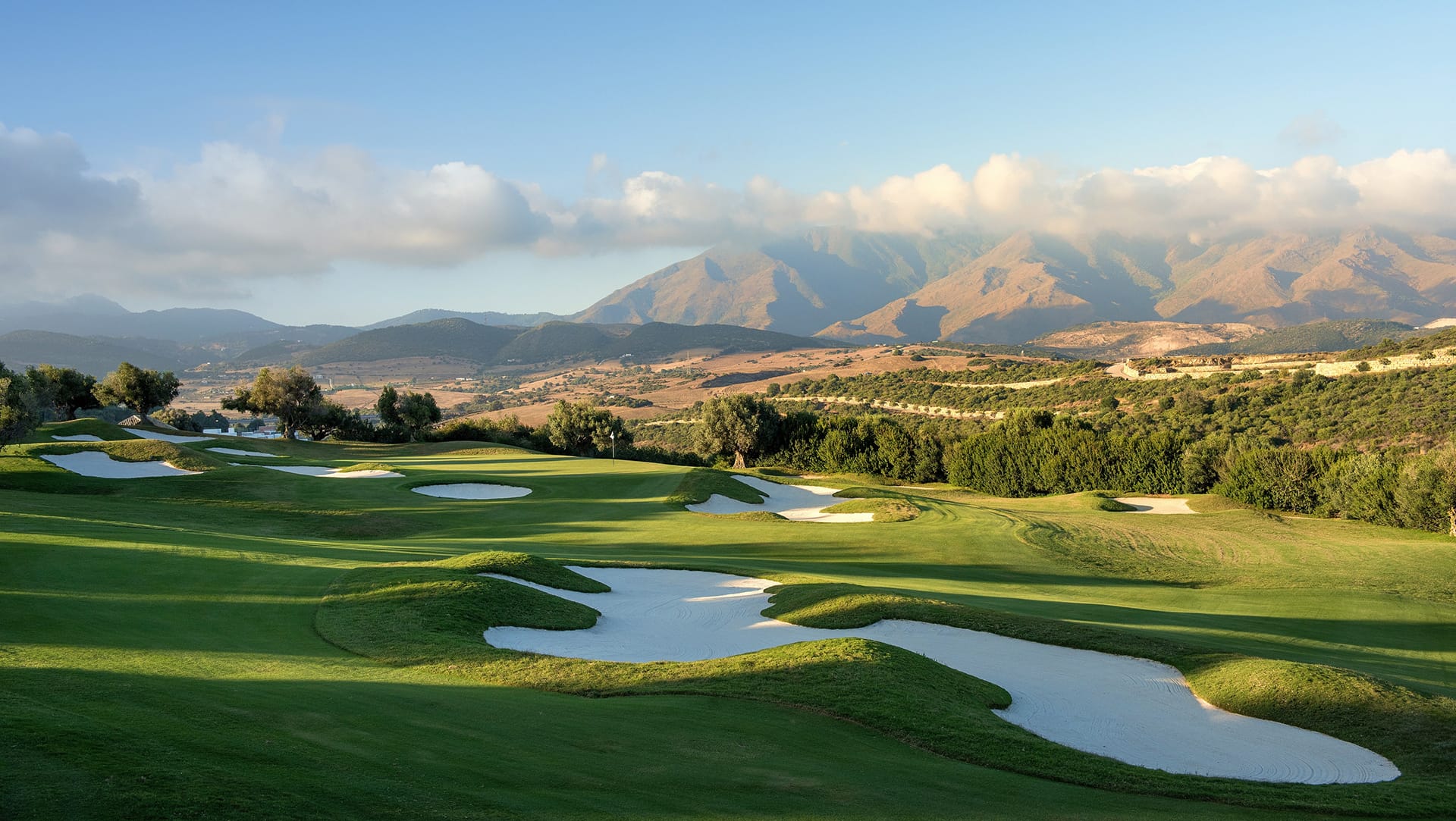 Scenic view of a golf course with sand traps and lush greenery, backdrop of mountains under a blue sky with clouds.