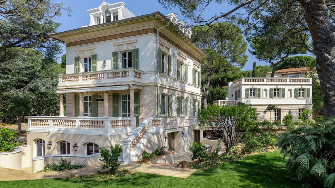 Elegant three-story white villa with decorative balconies surrounded by lush greenery and trees under a clear blue sky.