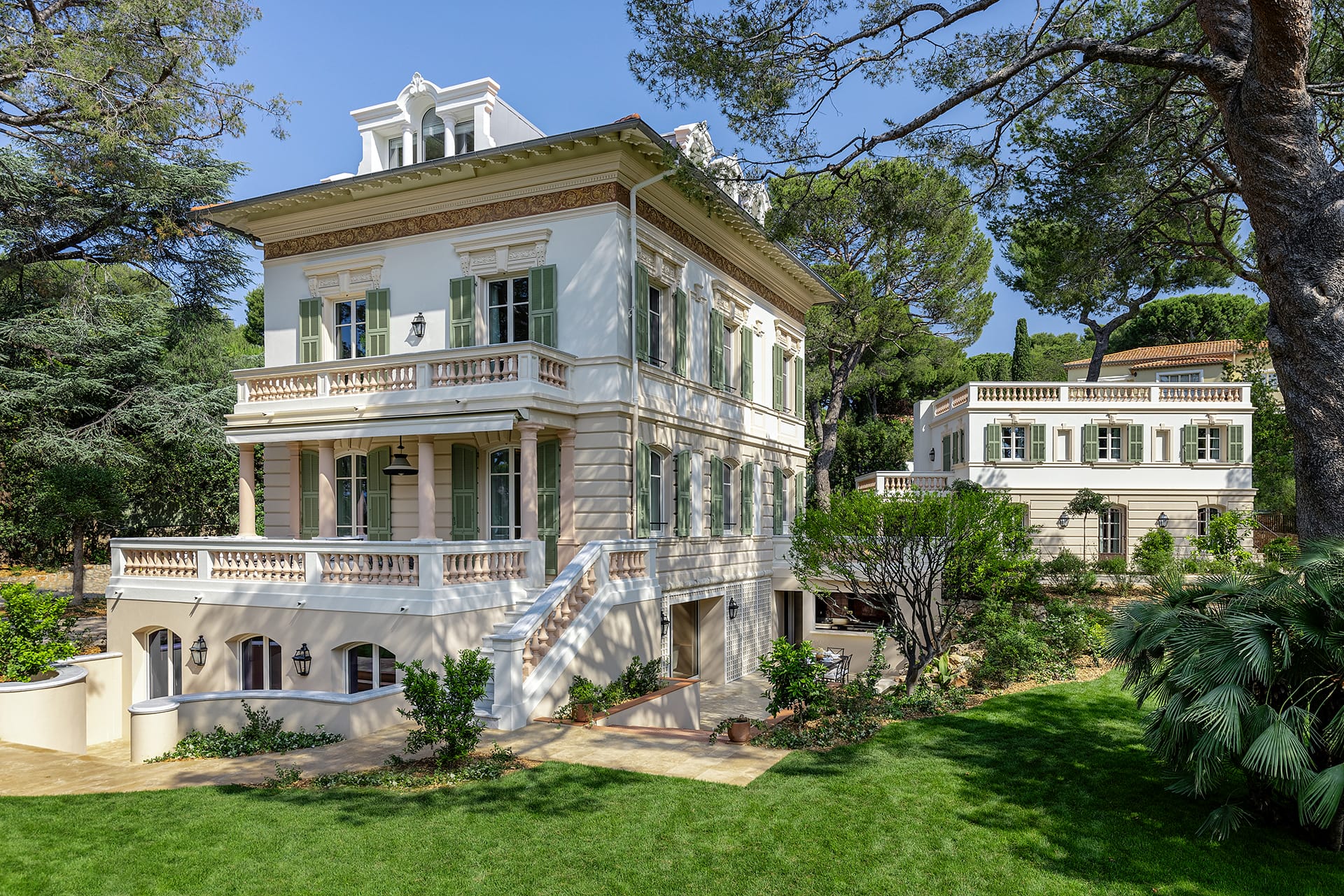 Elegant three-story white villa with decorative balconies surrounded by lush greenery and trees under a clear blue sky.