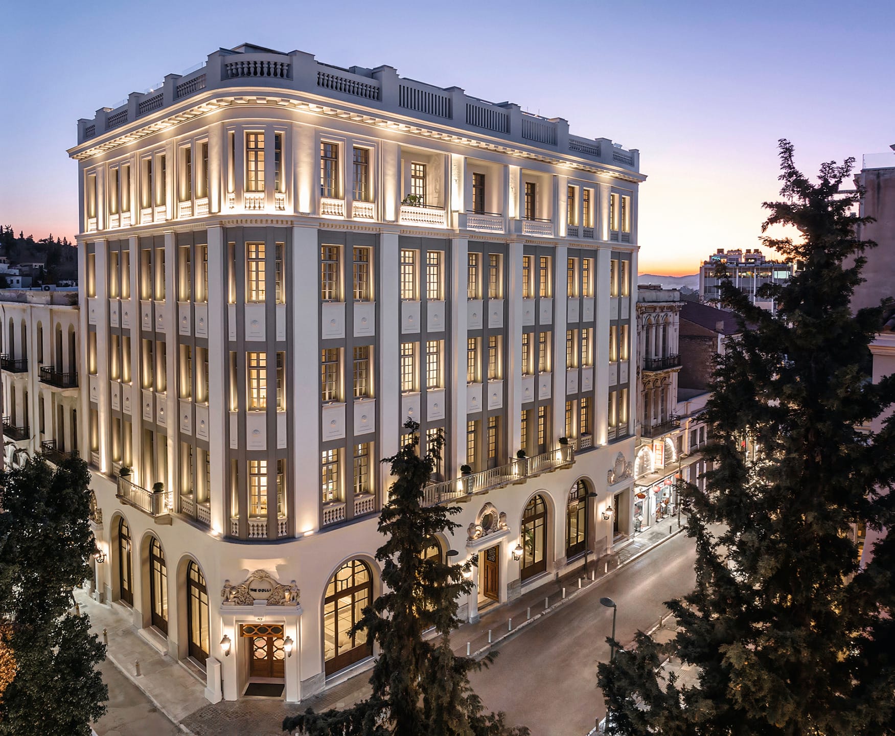 Historic multi-story building at dusk with illuminated windows and a classic facade surrounded by trees.