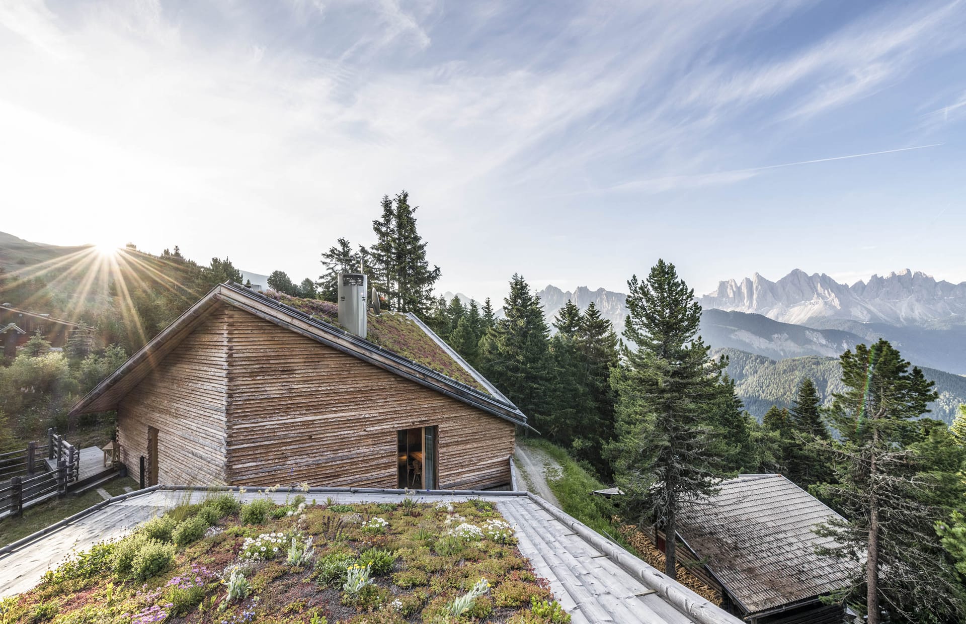 Rustic cabin with a green roof surrounded by pine trees and mountains under a clear sky with the sun peeking through.