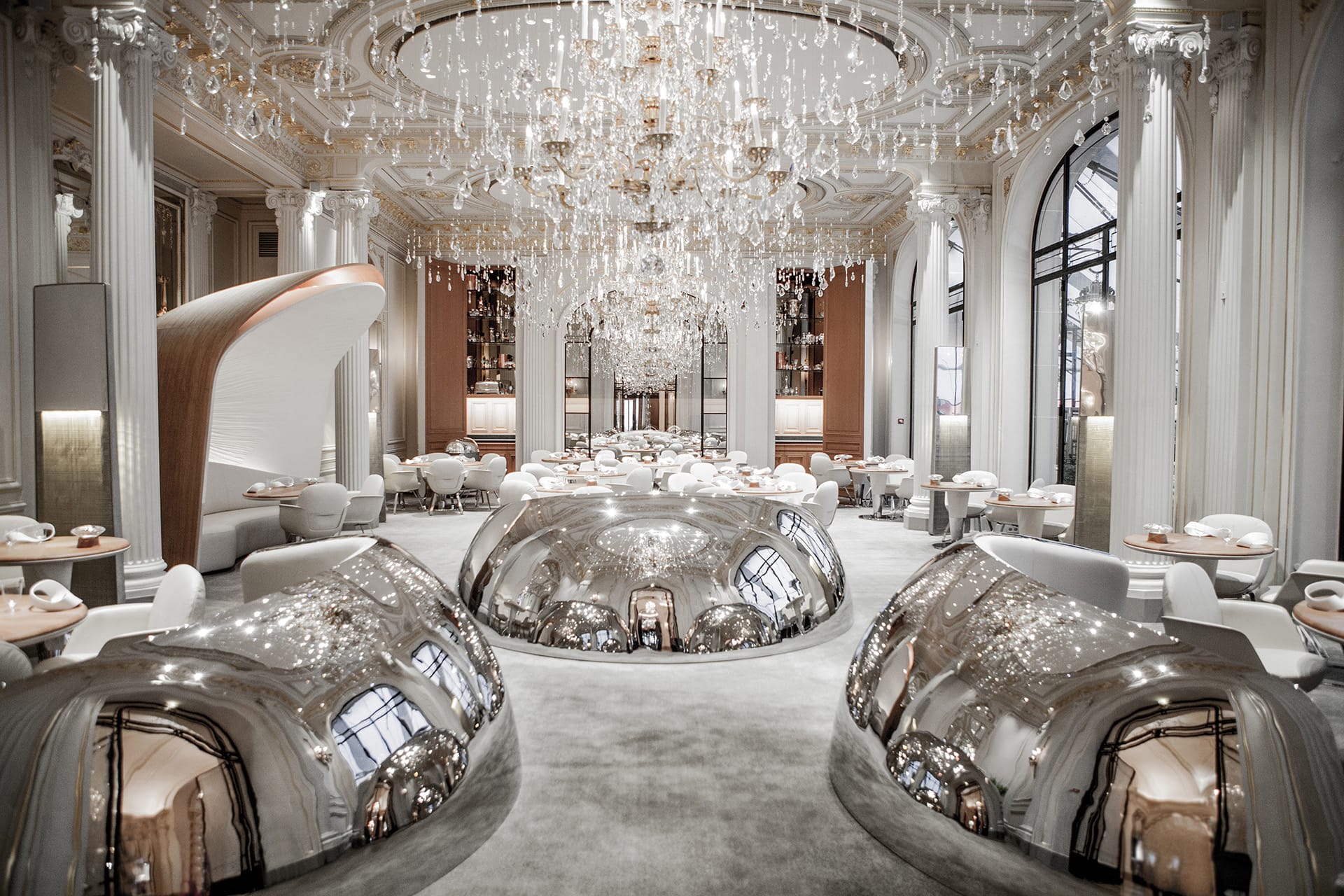 Elegant restaurant interior with chandeliers, luxurious decor, white seating, and reflective metallic sculptures
