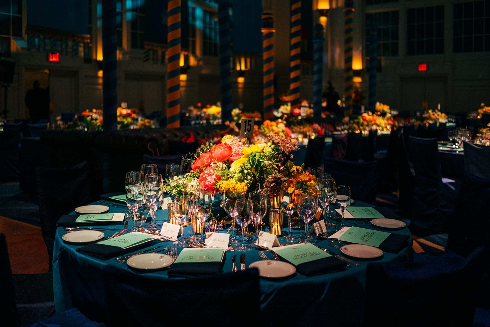 Elegant banquet table set with glassware, colorful floral centerpiece, and dim, ambient evening lighting in a grand hall.