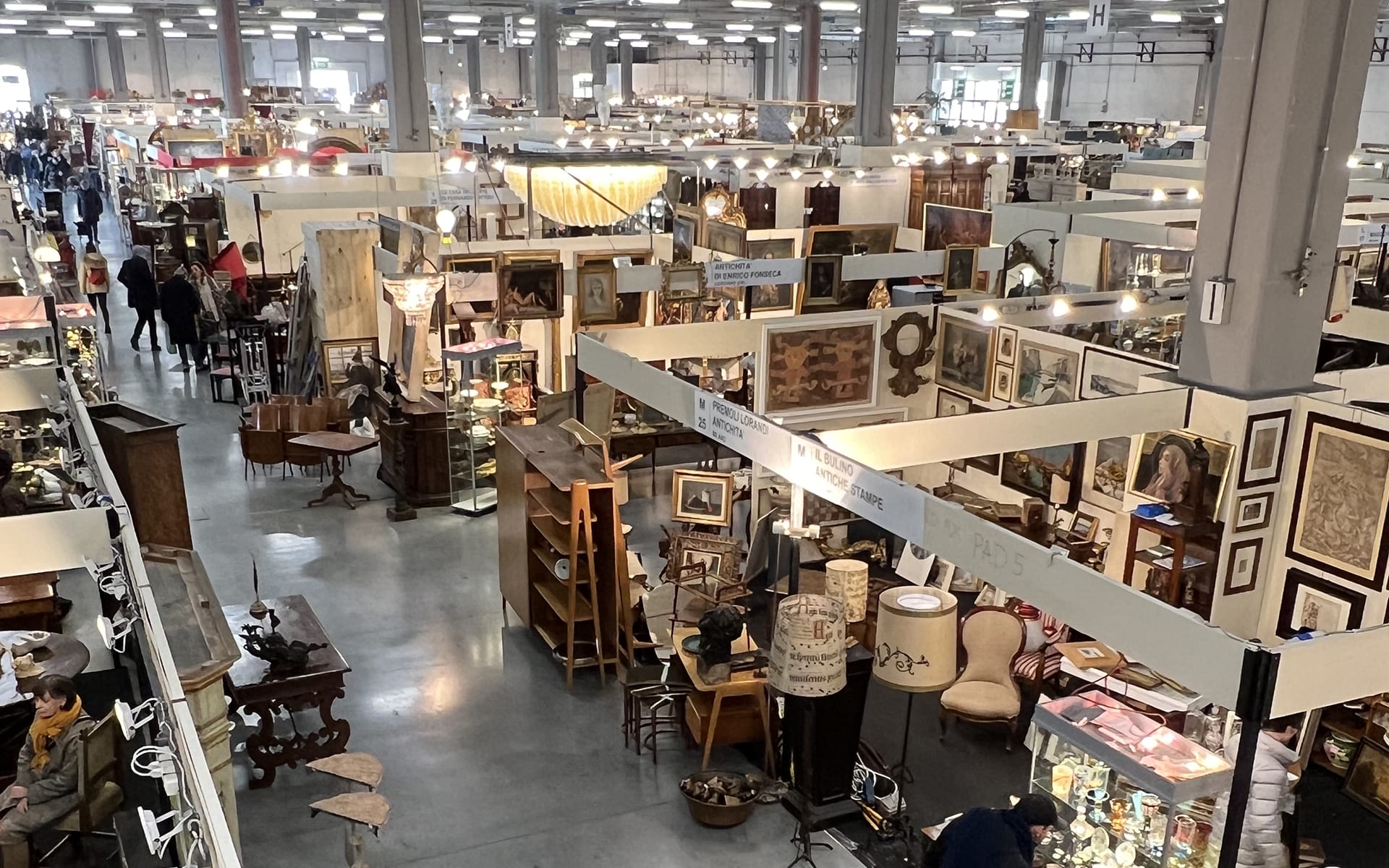 A large indoor antique market with various stalls displaying vintage furniture, artwork, and collectibles under bright lights.
