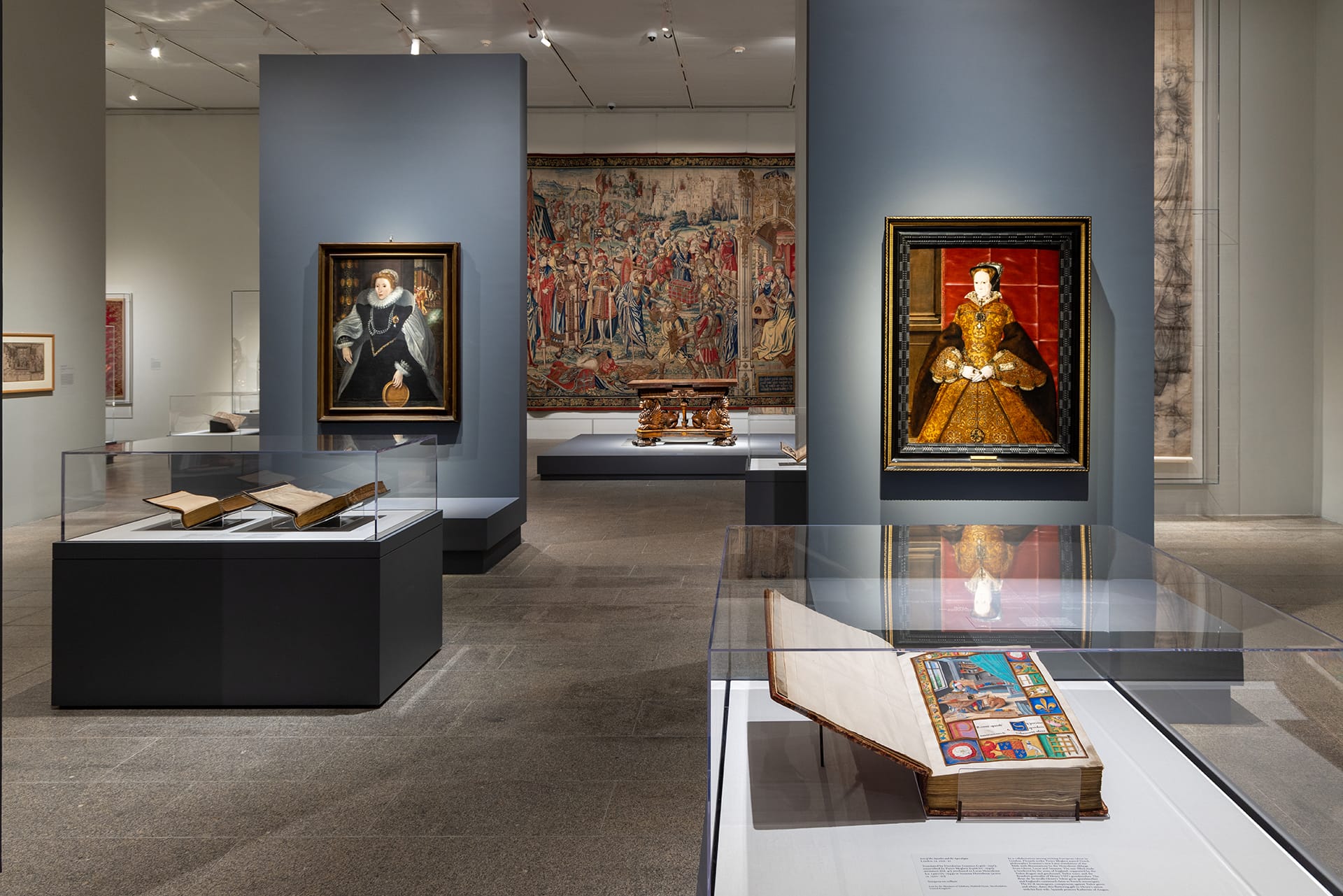 Art museum gallery with framed paintings, large tapestry, and display cases featuring open historical books on stands.