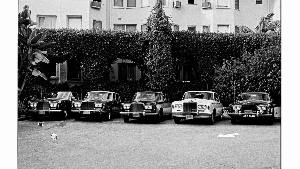 Black and white photo of six vintage cars parked in a row against a background of foliage and a building.