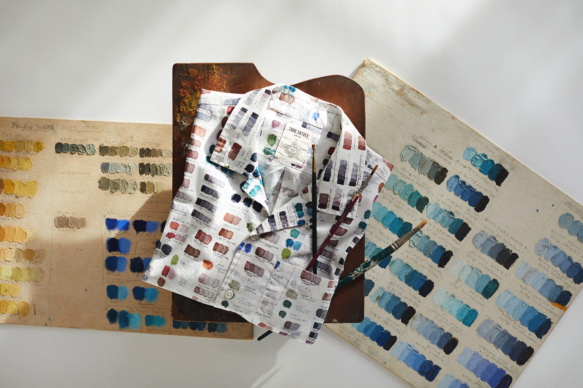 Colorful shirt with paint swatch patterns displayed on a wooden table with paint palettes and brushes.