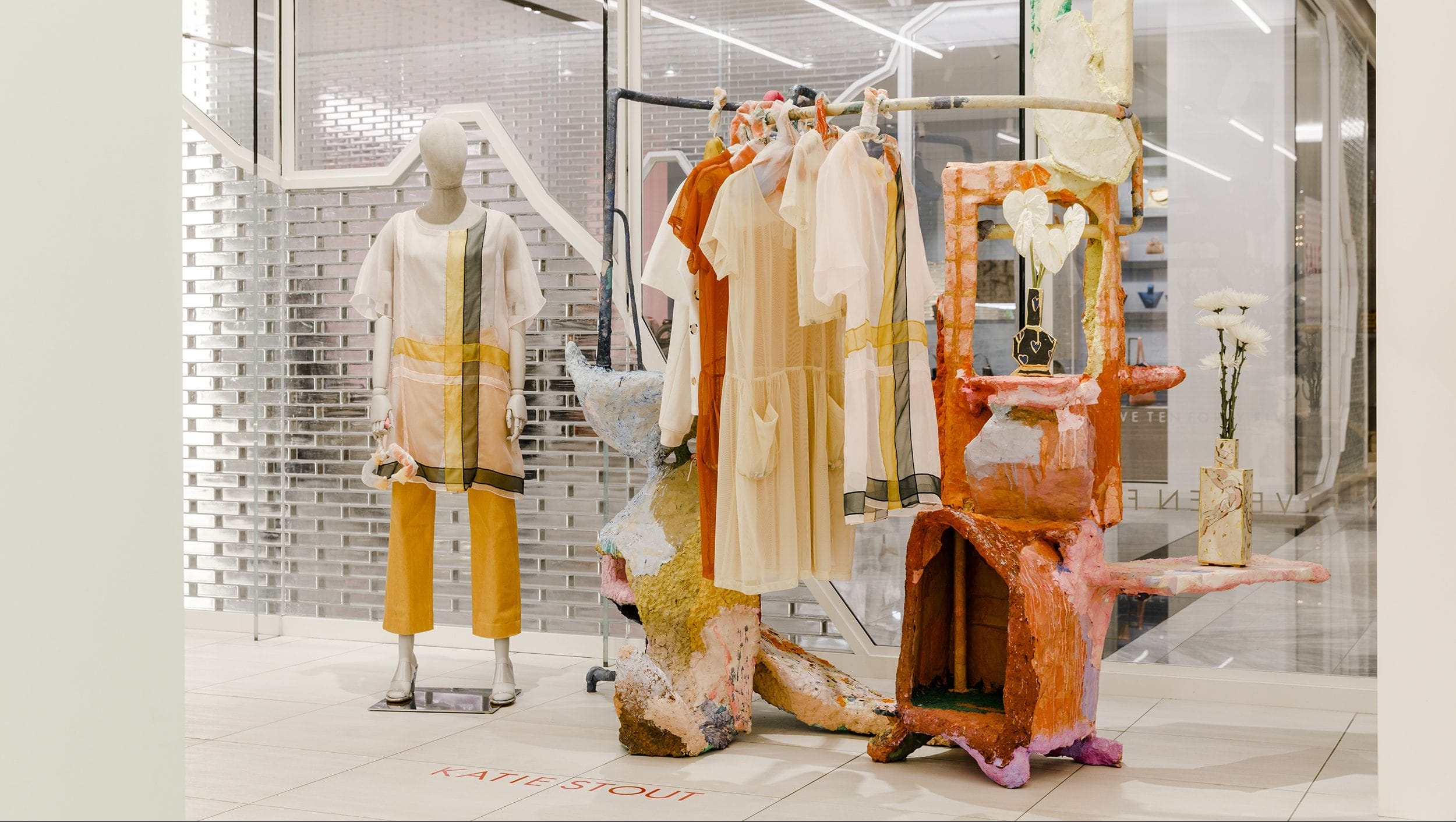 Store display with a mannequin in a stylish yellow and white outfit beside vibrant, artistic fixtures and hanging clothes.