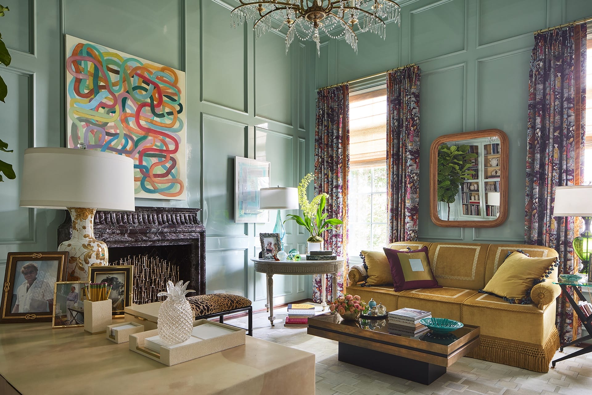 Elegant living room with teal walls, patterned curtains, a gold sofa, vibrant art, and a chandelier. Warm, inviting ambiance.