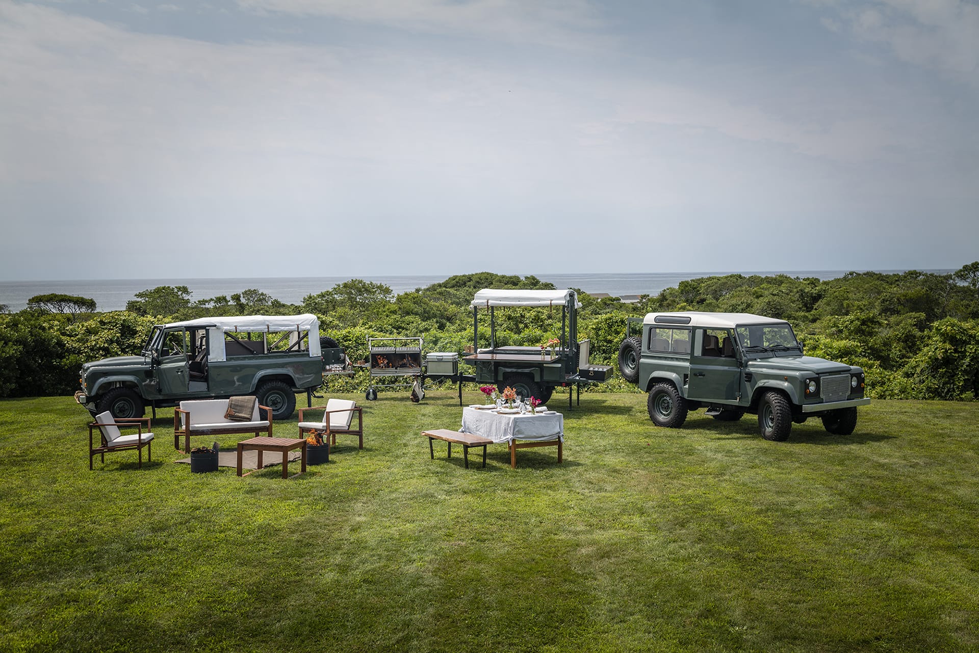Outdoor gathering setup with two vintage off-road vehicles, a trailer, and picnic arrangements on a grassy field near the sea.