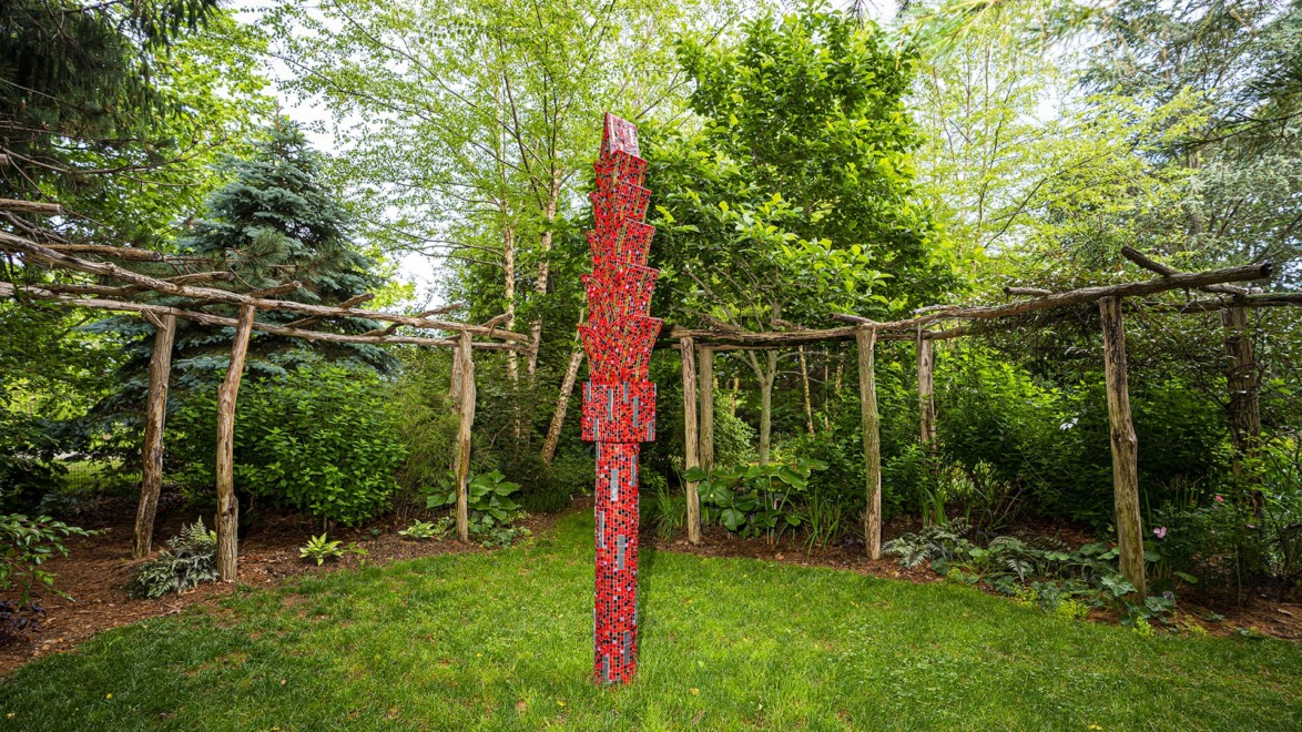 Red abstract sculpture stands vertically in a lush green garden surrounded by trees and rustic wooden archways.