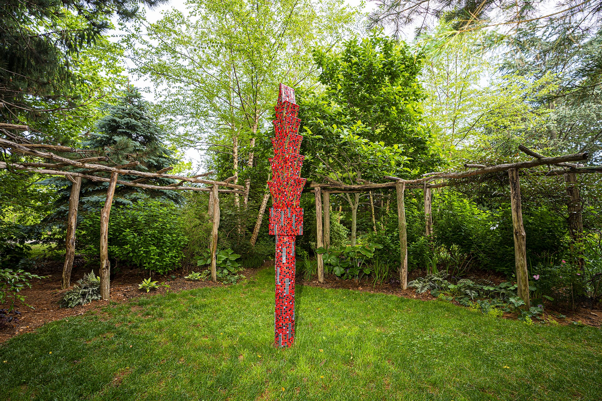 Red abstract sculpture stands vertically in a lush green garden surrounded by trees and rustic wooden archways.