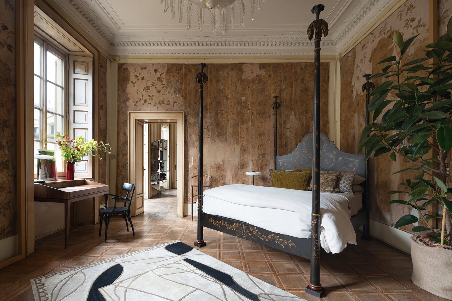 Elegant bedroom with a four-poster bed, ornate ceiling, patterned wallpaper, wooden floor, and large window with a desk nearby.