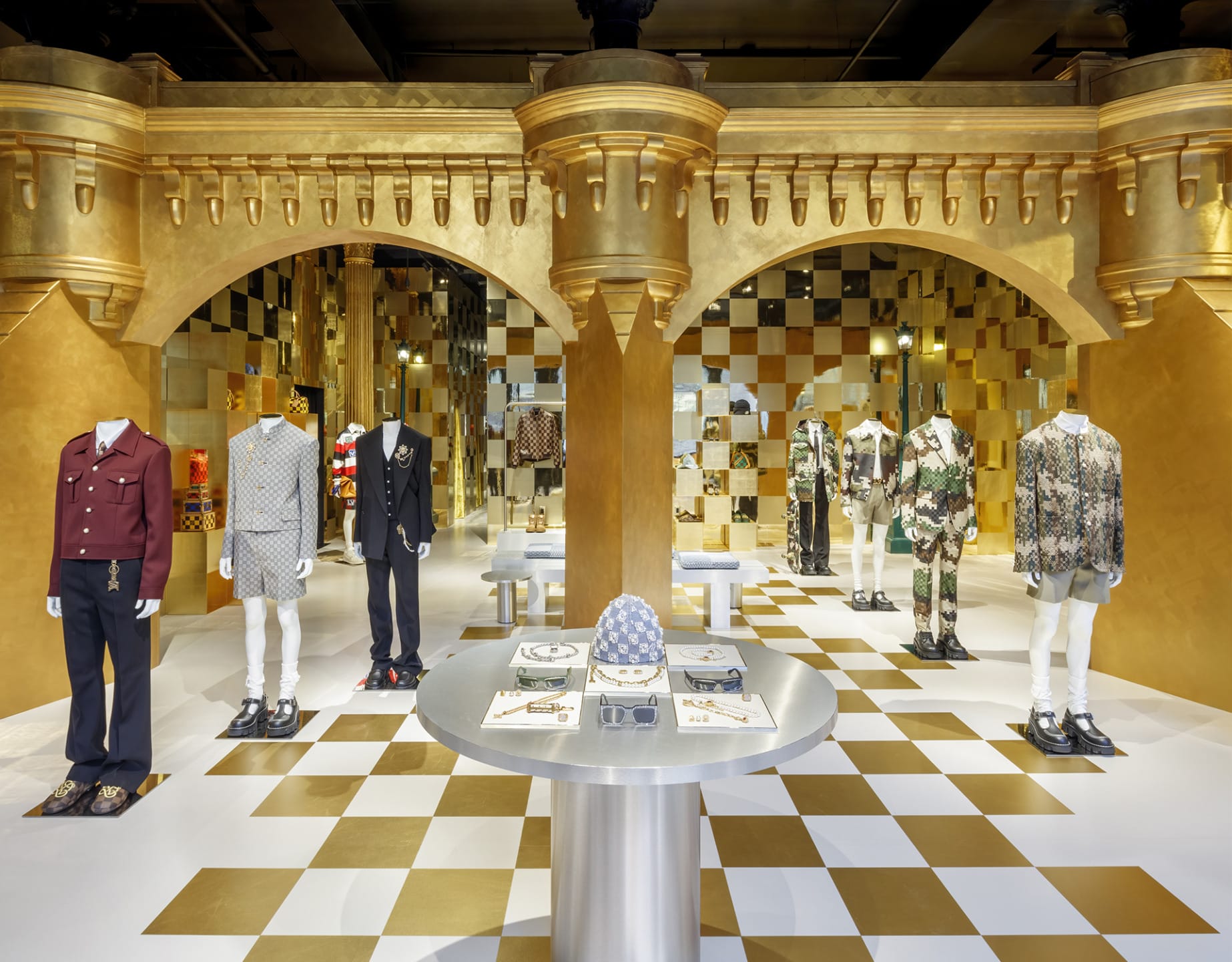 Luxury fashion store interior with gold decor, mannequins in designer clothing, and a display table with accessories.