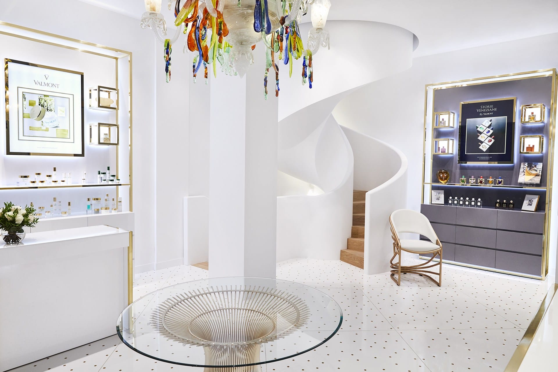Modern, elegant interior with spiral staircase, colorful chandelier, and luxury product displays on sleek, white surfaces.