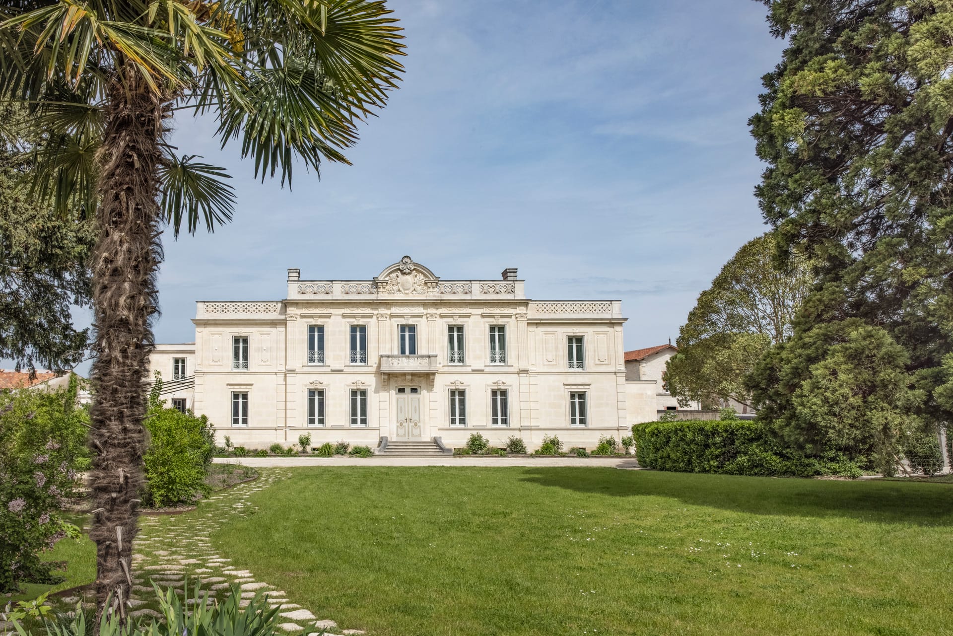 Elegant white mansion with large windows, surrounded by lush greenery and palm trees on a sunny day.