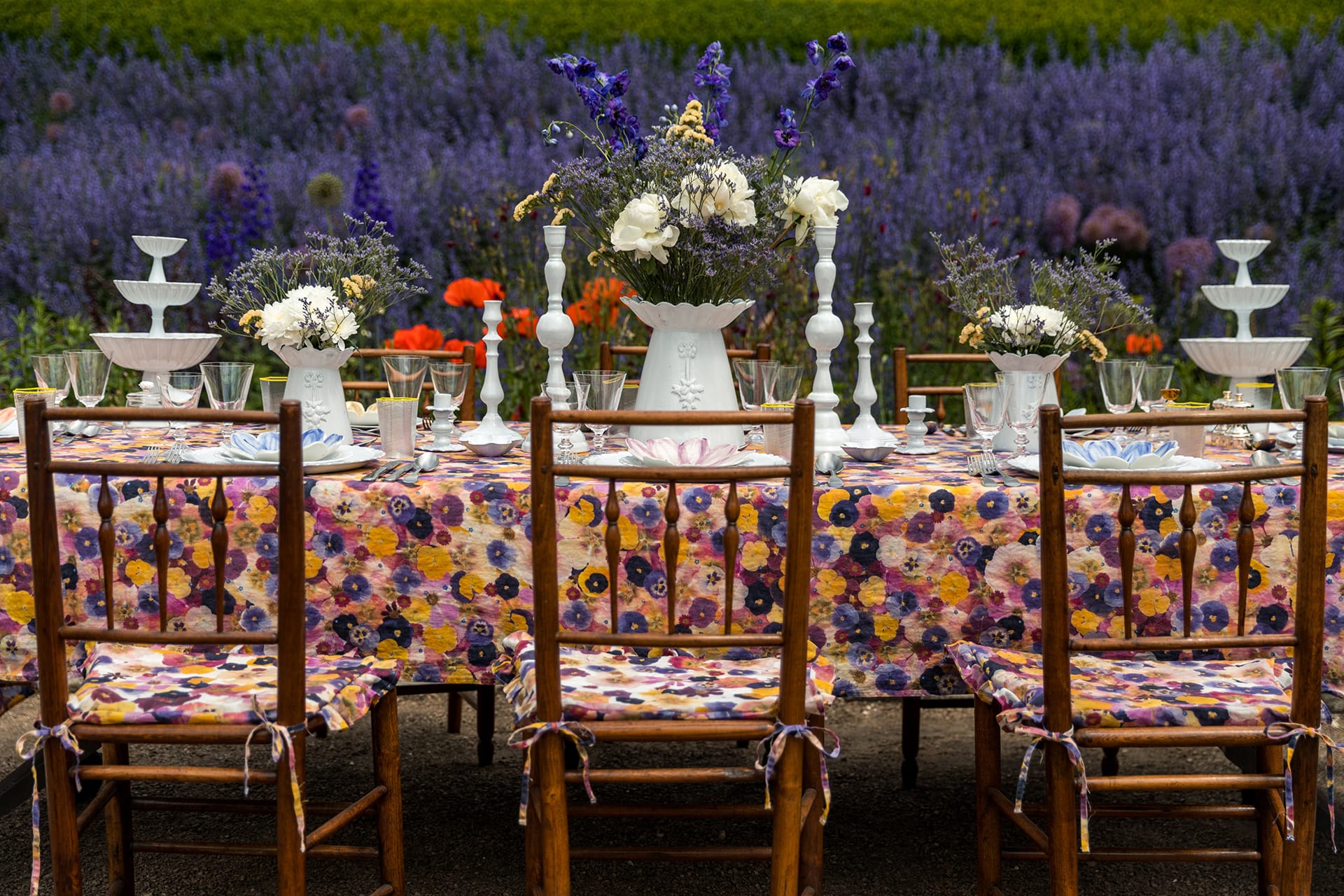 Colorful outdoor dining table set with floral tablecloth, rustic chairs, and white vases filled with flowers by a lavender field.