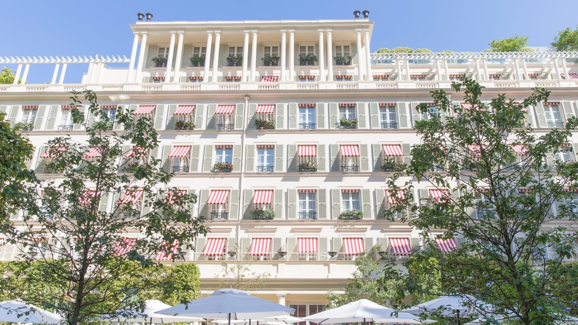 Historic white building with red-striped awnings, surrounded by lush trees, under a clear blue sky.