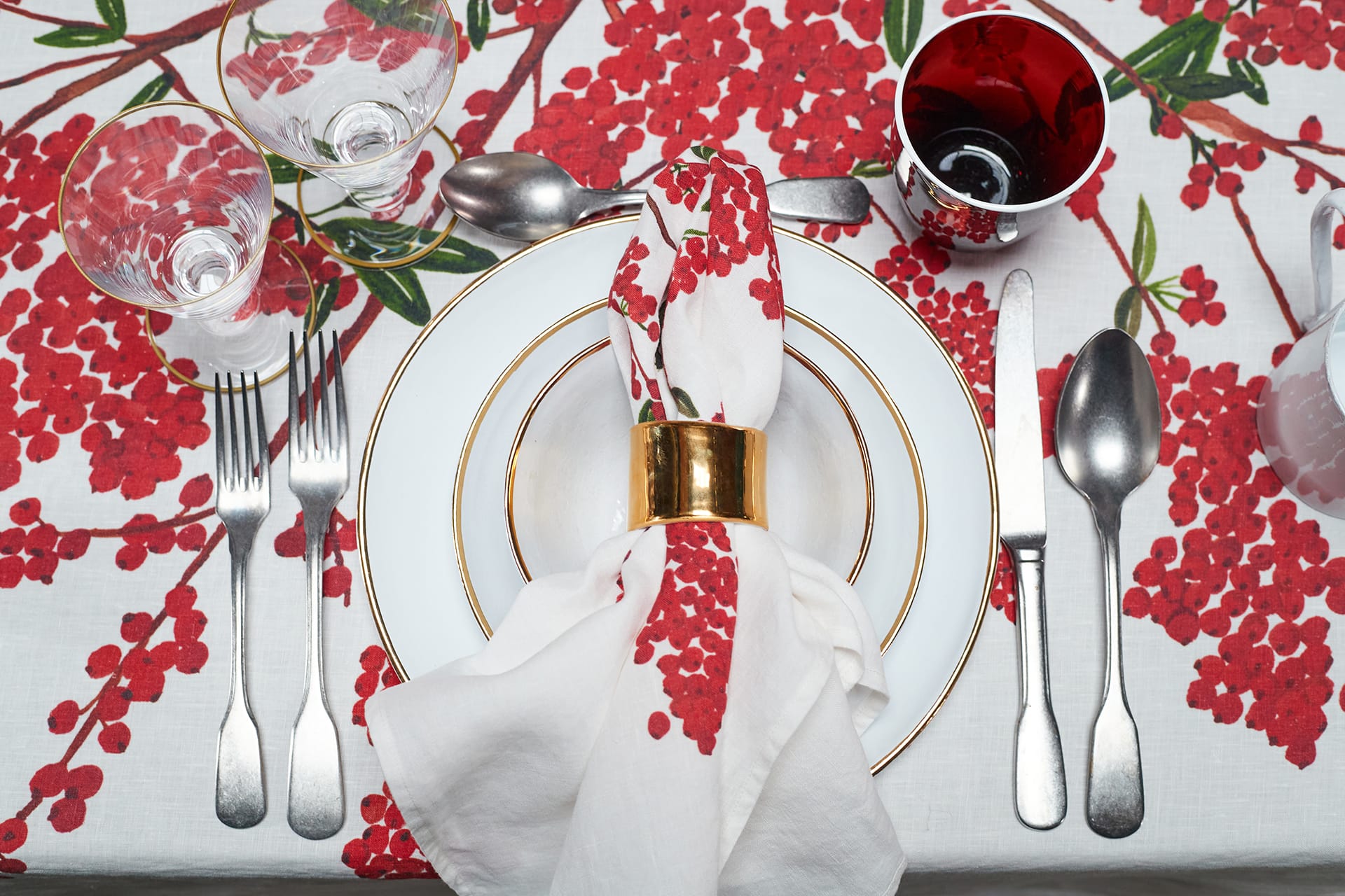 Festive table setting with white plates, gold accents, and red berry-patterned tablecloth, complete with glassware and cutlery.
