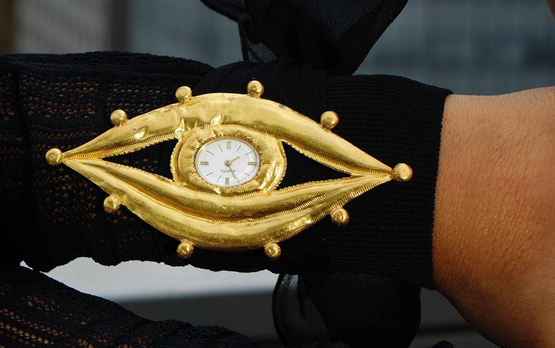 Gold eye-shaped wristwatch with black lace gloves, showing a white watch face.