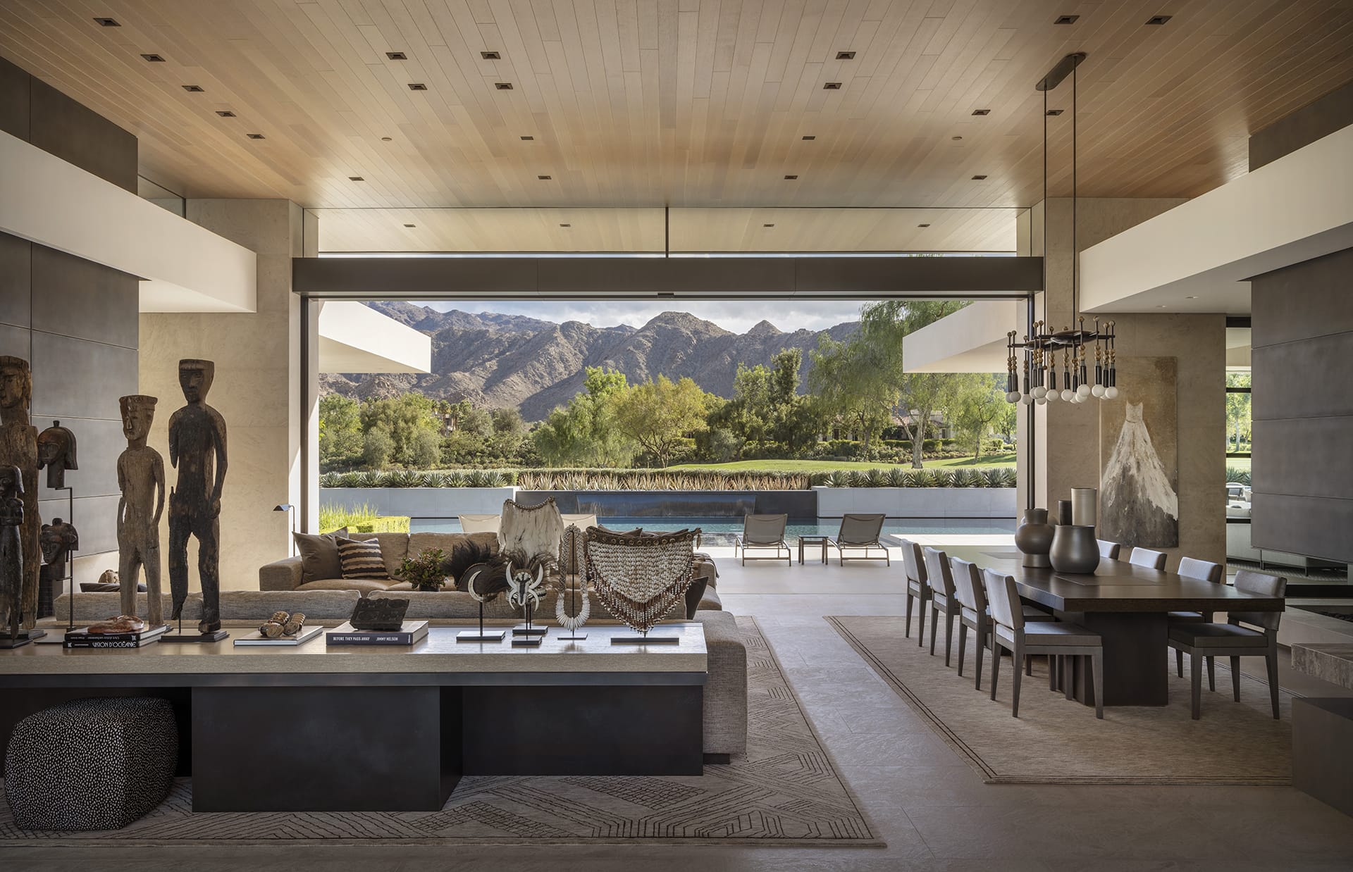 Modern open-concept living room with large glass windows, mountain views, and stylish decor featuring sculptures and a long dining table.