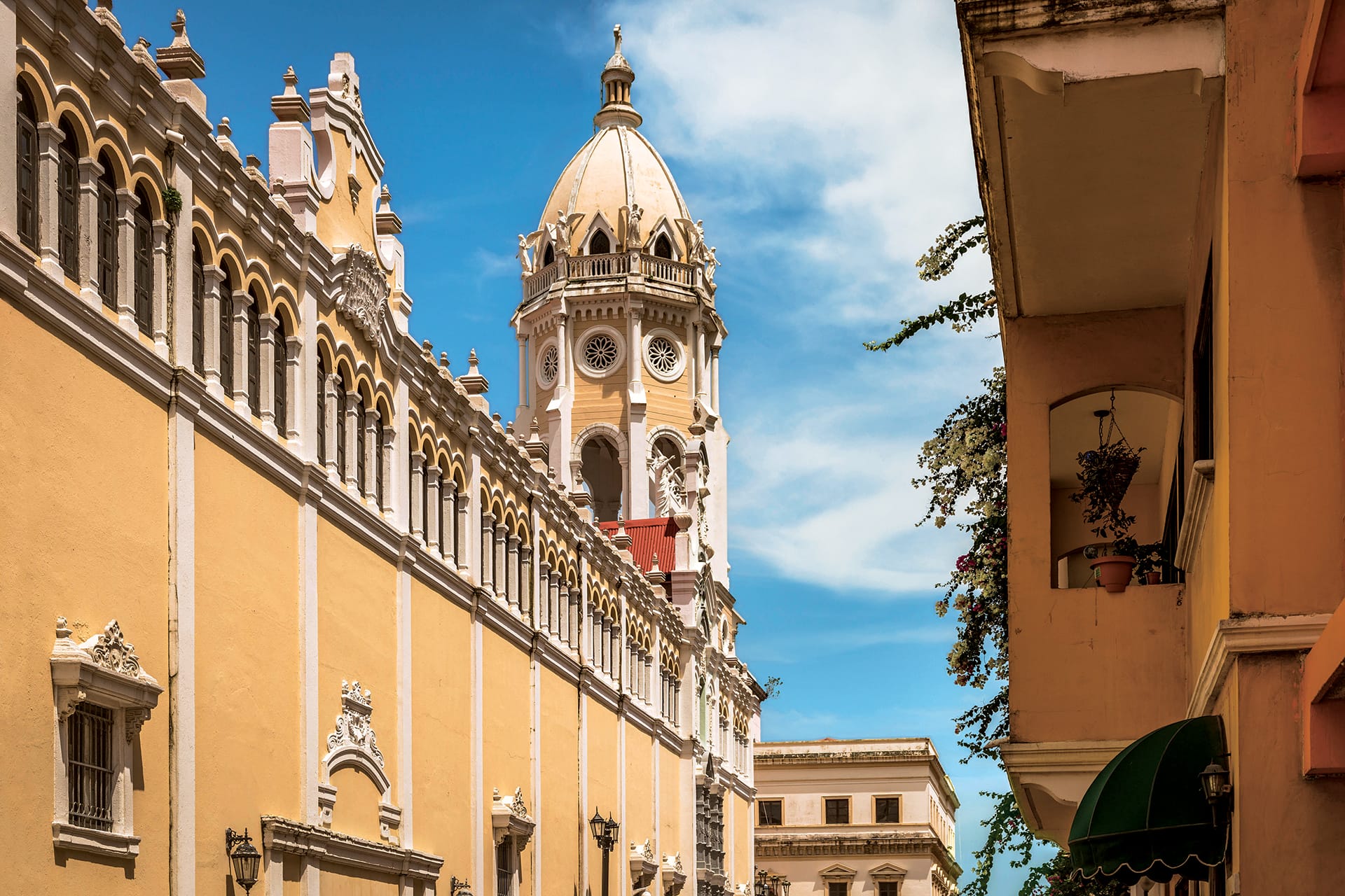 Historic yellow building with ornate tower and clock against a blue sky in a cityscape with adjacent buildings.